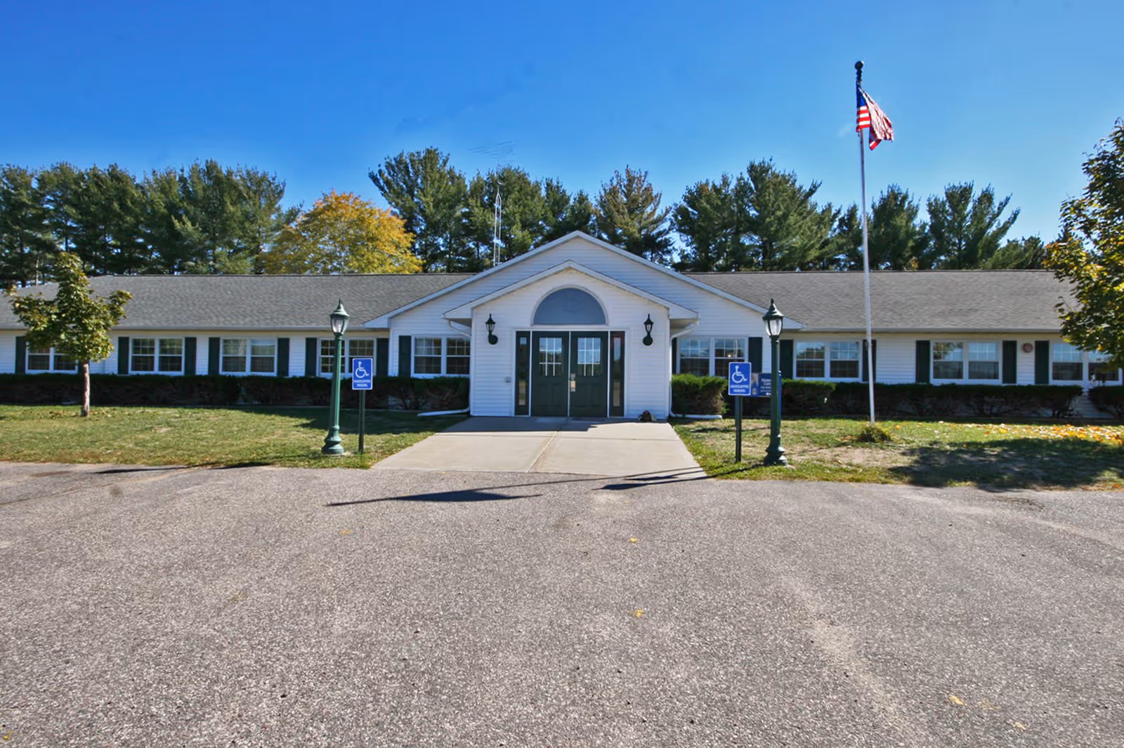Front exterior view of a single-story senior living facility building with white siding and green doors. There are two handicap parking signs near the entrance, two lamp posts, an American flag on a flagpole to the right, and trees in the background under a clear blue sky.