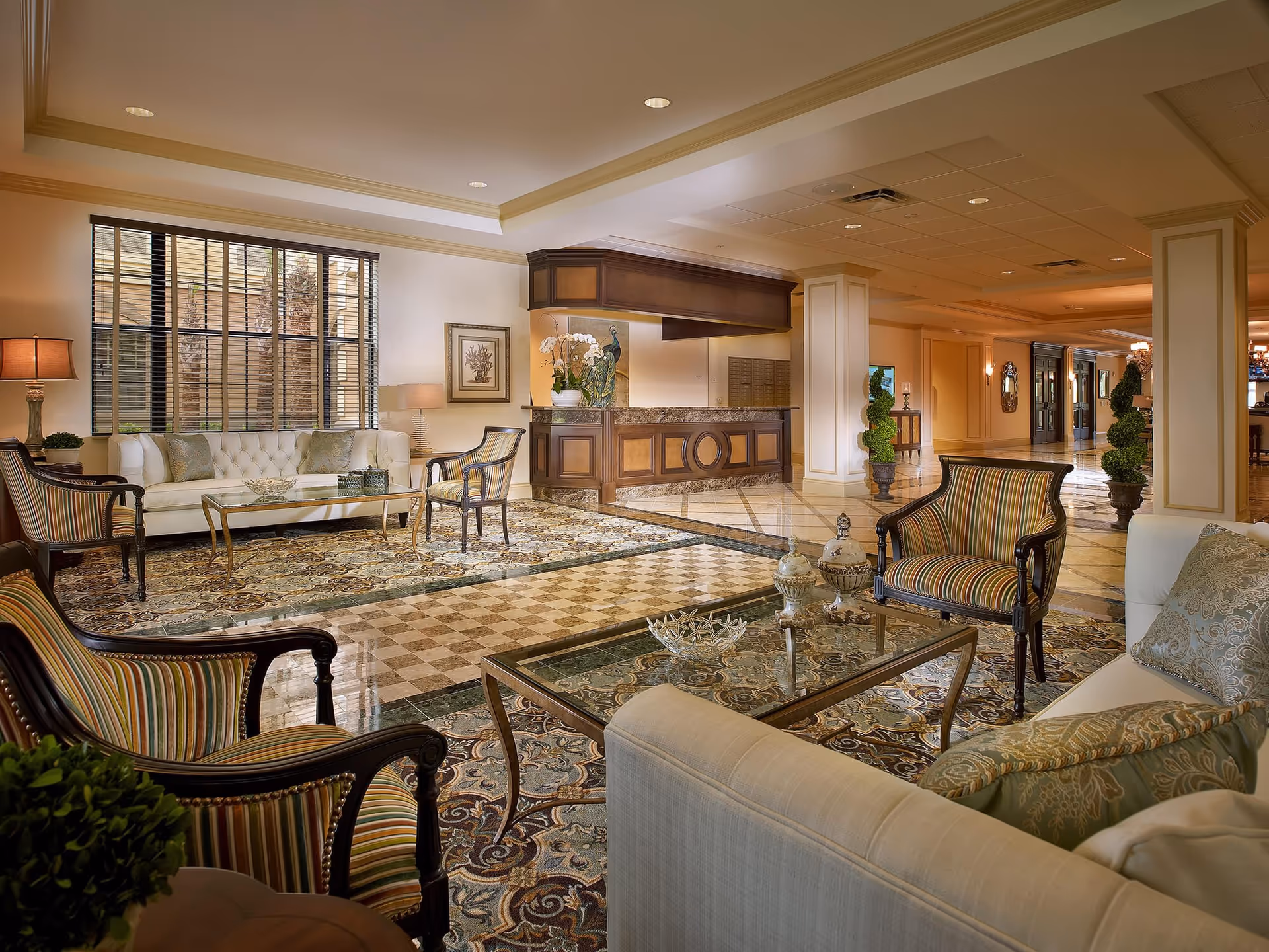 A spacious and elegant senior living facility lobby with comfortable seating including striped armchairs and white sofas arranged around glass coffee tables on patterned rugs. The area features polished marble floors, a reception desk with wood paneling, decorative plants, and warm lighting from lamps and ceiling fixtures.