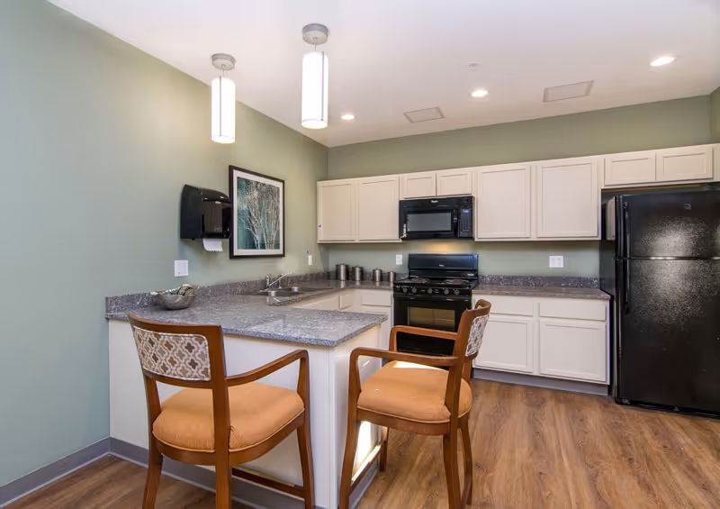 A small kitchen area with light green walls, white cabinets, a black refrigerator, black microwave, and black stove. There is a gray countertop with a sink and two wooden chairs with orange cushions and patterned backs. Two pendant lights hang from the ceiling, and a framed picture is on the wall above the countertop.