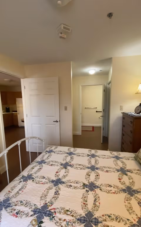 A bedroom with a bed covered in a quilt featuring a floral and geometric pattern. To the right is a wooden dresser with a lamp and a clock on top. The room has beige walls and an open door leading to a hallway and another room. The kitchen is partially visible through an open door on the left.