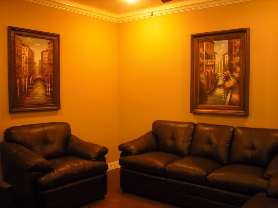 A cozy seating area with dark leather armchairs and a sofa beneath framed paintings on warm yellow walls.