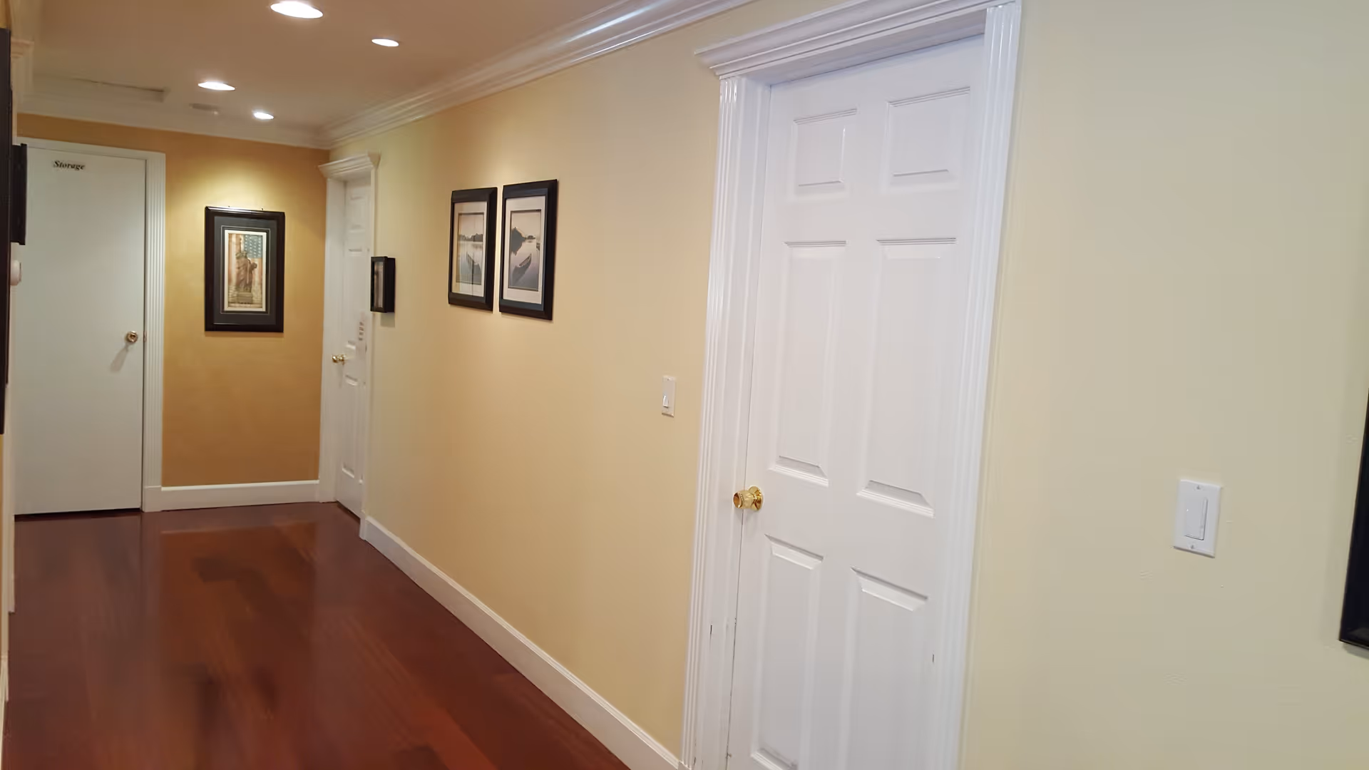 Well-lit interior hallway with hardwood floors, beige walls, white doors, and framed wall art.