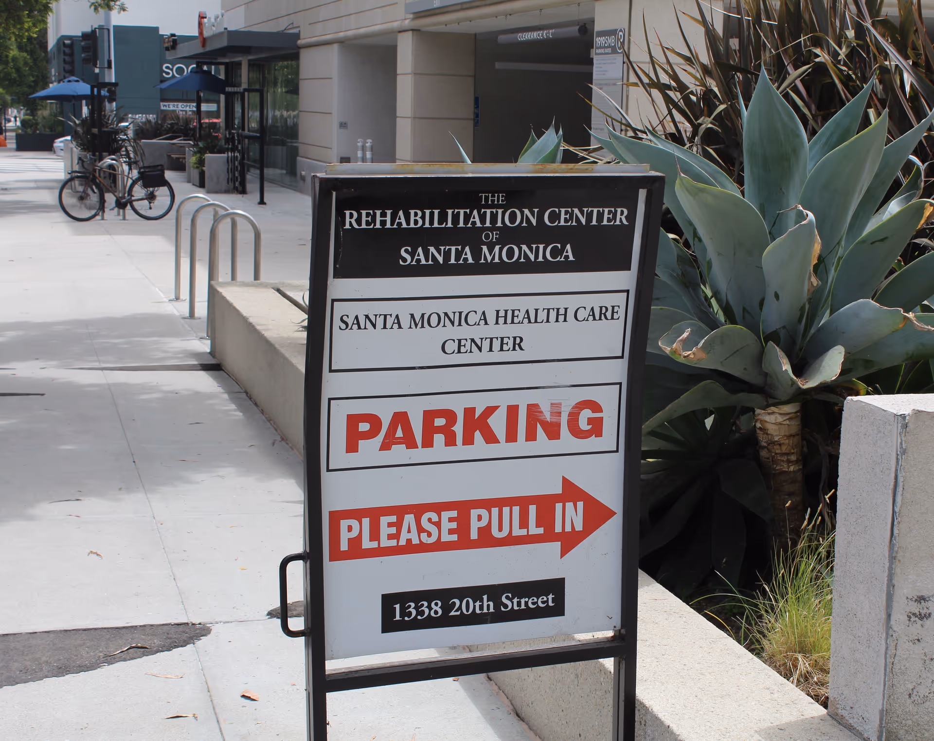 A sidewalk sign outside a building with text indicating it is The Rehabilitation Center of Santa Monica and Santa Monica Health Care Center. The sign directs parking with the message 'PARKING PLEASE PULL IN' and provides the address 1338 20th Street. There are plants and a bicycle rack nearby on the sidewalk.