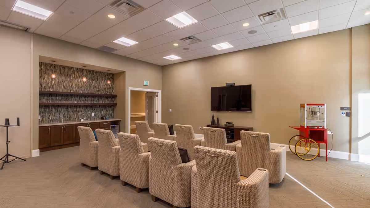 A cozy media room with two rows of beige upholstered armchairs facing a wall-mounted flat-screen TV. To the left, there is a small kitchenette area with a mosaic tile backsplash, wooden cabinets, and shelves. On the right side of the room, there is a red and yellow popcorn machine on wheels. The room has beige walls, a carpeted floor, and a drop ceiling with recessed lighting.