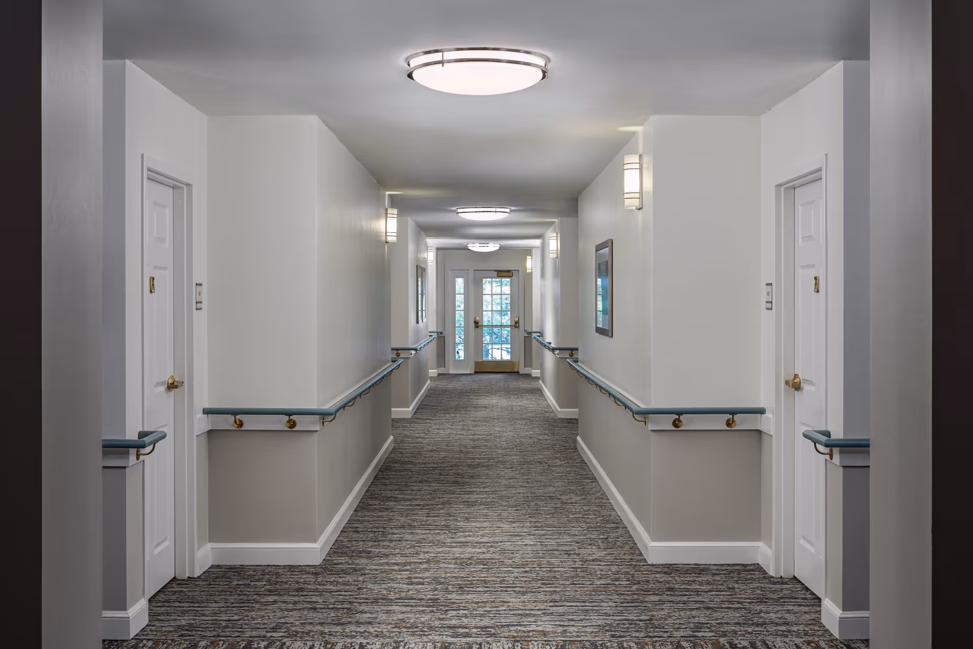Carpeted hallway in a senior living facility with handrails, white doors, and ceiling lights leading to a glass exit door.