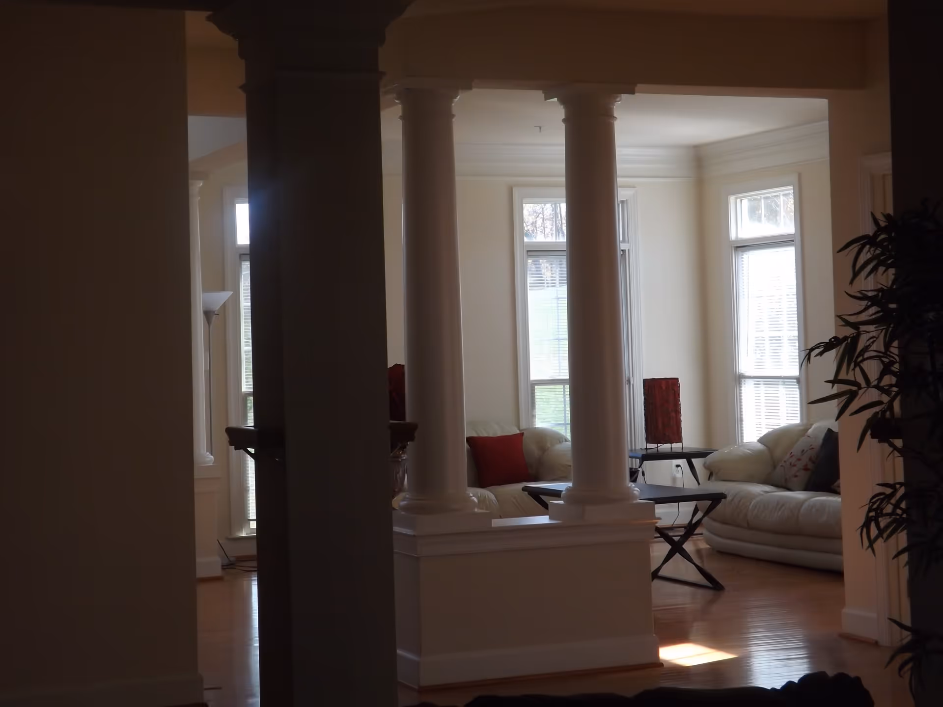 Interior view of a living room area with white leather sofas, red and patterned cushions, a small black coffee table, tall white columns, large windows with blinds, and wooden flooring. There is a floor lamp and a plant partially visible on the right side.