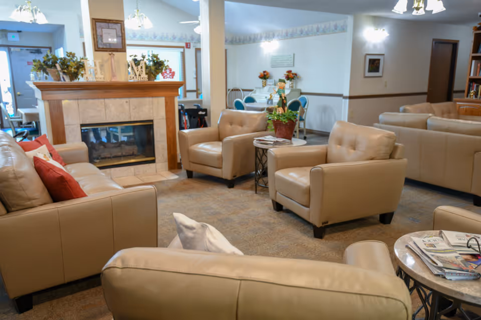 A cozy living room area in Havelock Manor featuring beige leather sofas and armchairs arranged around a fireplace with a wooden mantel. The room is decorated with plants, flowers, and framed pictures, with a small round table holding magazines and a potted plant. In the background, there is a dining area with blue chairs and a white piano.