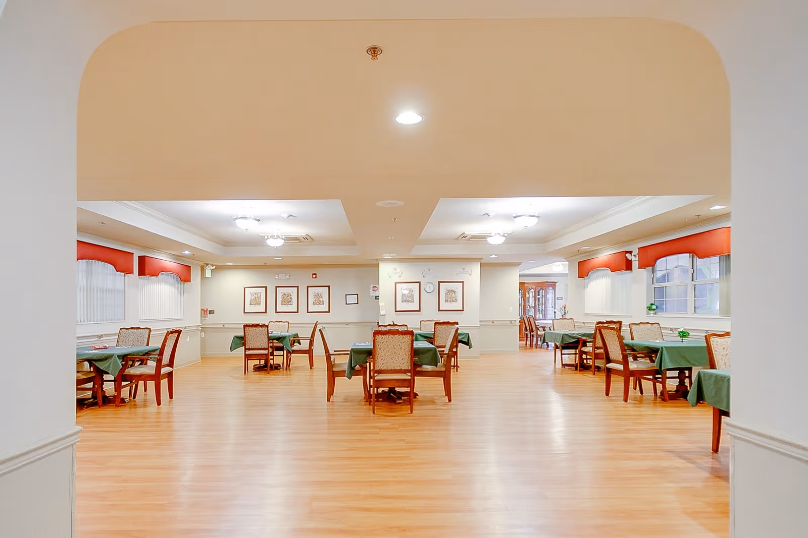 A spacious dining room in a senior living facility with wooden floors, several tables covered with green tablecloths, and chairs arranged around them. The room has soft lighting, framed artwork on the walls, and windows with red valances.