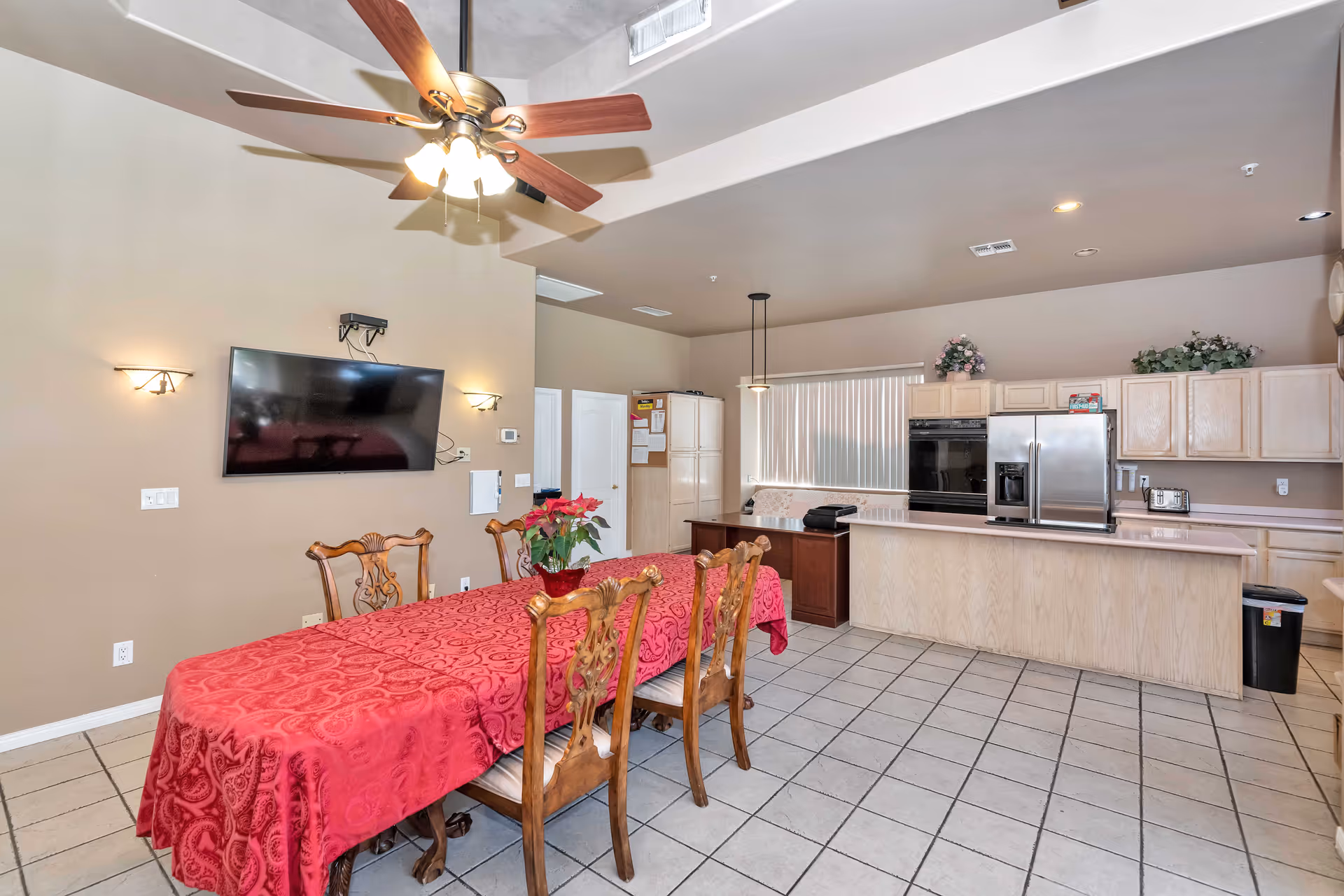 A spacious dining area with a long wooden table covered by a red patterned tablecloth and six ornate wooden chairs. Above the table is a ceiling fan with lights. On the wall, there is a flat-screen TV mounted between two wall sconces. The dining area opens into a kitchen with light wood cabinets, a large island, stainless steel refrigerator, and built-in oven. The floor is tiled, and there are decorative plants on top of the kitchen cabinets.