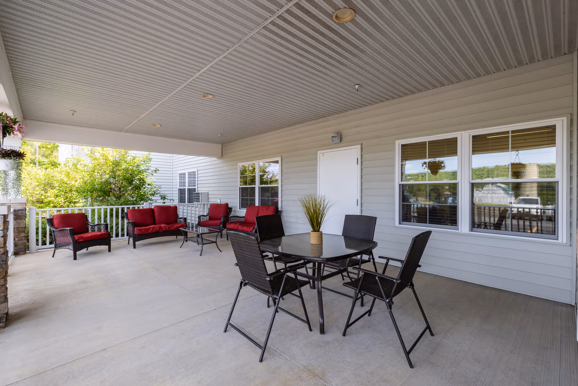 Covered outdoor patio area with a seating arrangement including a glass-top table with four black chairs and a small plant centerpiece. There are also two red cushioned armchairs and a red cushioned loveseat against the wall. The patio has a light gray ceiling and siding, with windows and a white door on the wall.