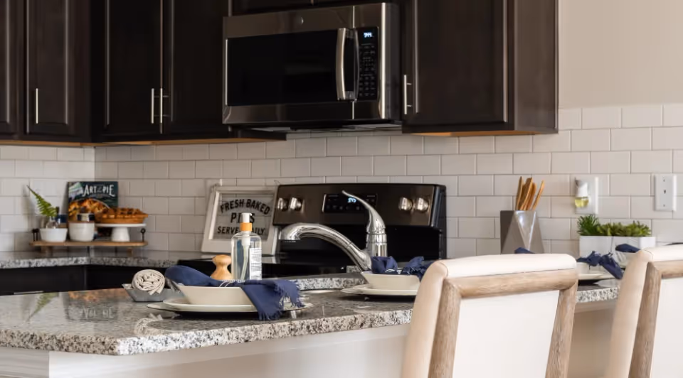 Modern kitchen with dark wood cabinets, a stainless steel microwave and stove, and a granite countertop island set with plates, napkins, and a bottle of hand soap. Decorative items include a sign that says 'Fresh Baked Pie Served Daily', a pie on a stand, and a knife block with utensils.