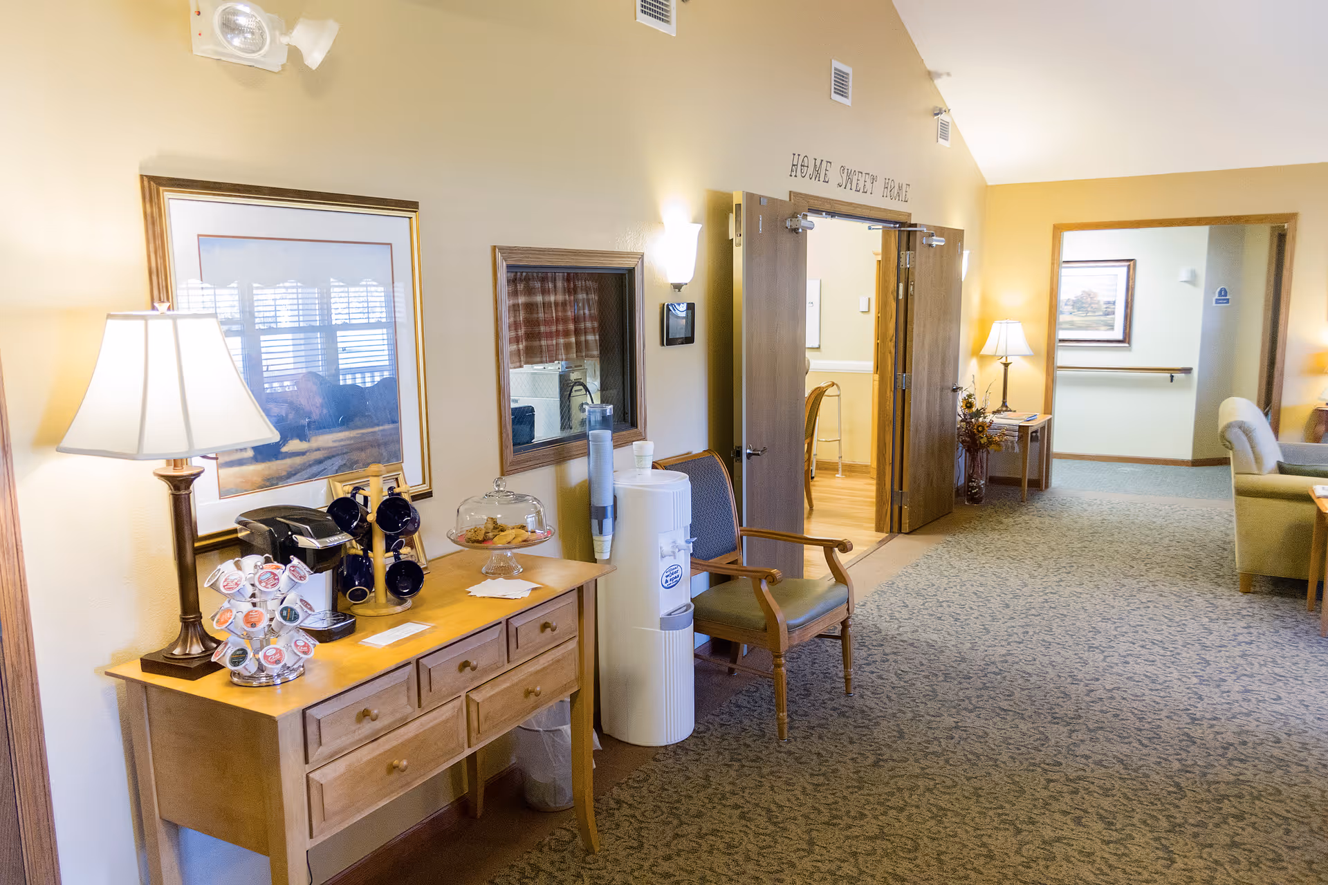 Interior hallway of a senior living facility with a wooden console table holding a lamp, coffee maker, coffee pods, and a glass dome with cookies. A water cooler and a wooden chair are next to the table. Double doors with the words 'Home Sweet Home' above them lead to another room. The hallway is carpeted and has warm yellow walls with framed artwork and additional seating visible in the background.