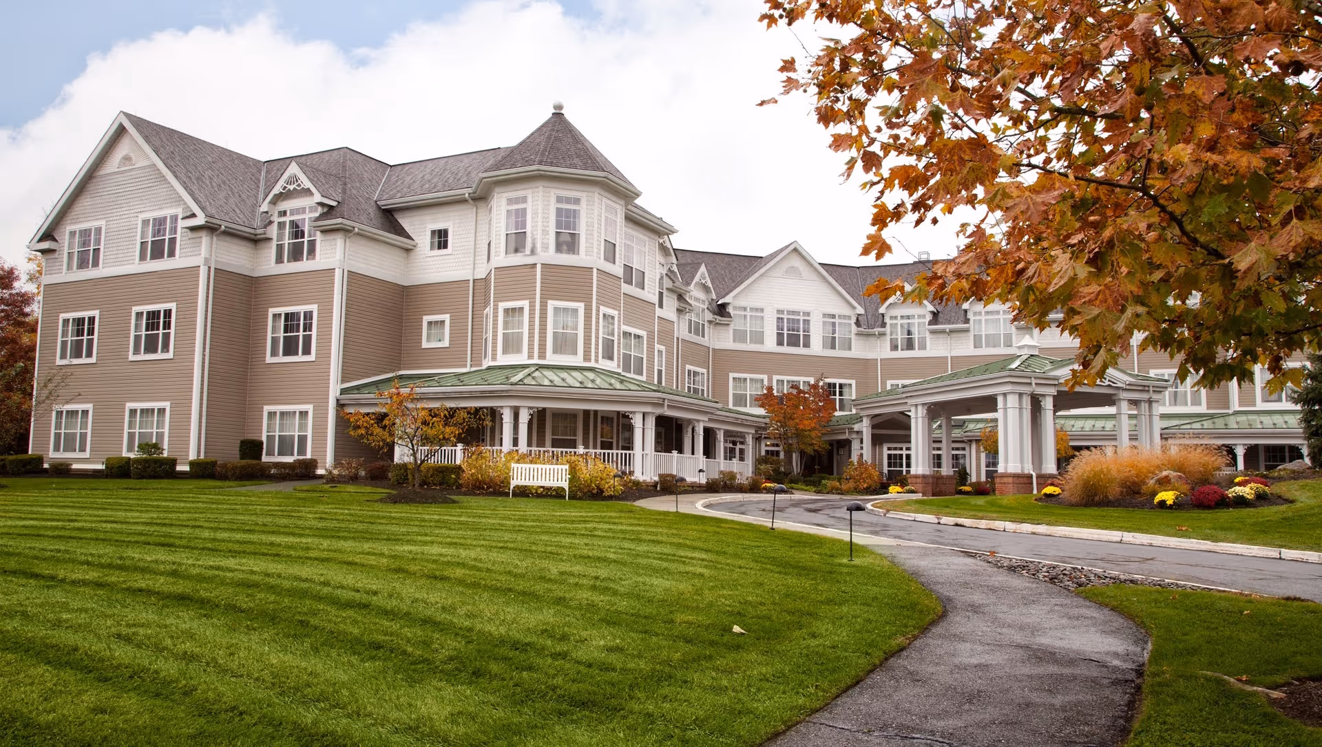 Front exterior of a large three-story senior living building with a circular driveway, manicured lawn, and autumn trees.