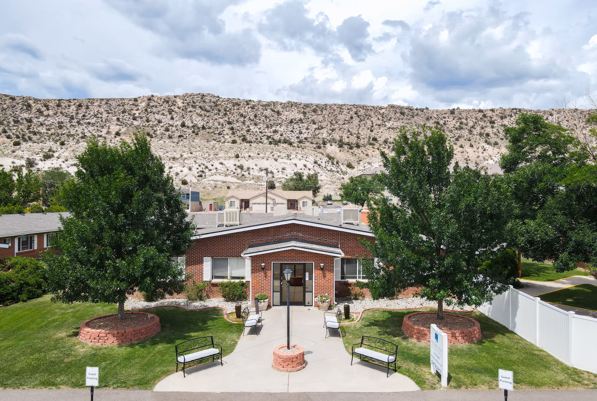 Front exterior view of Skyline Ridge Nursing and Rehabilitation Center, a single-story brick building with a central entrance, surrounded by green lawns, two large trees in circular brick planters, benches, and a lamppost. In the background, there is a rocky hillside under a partly cloudy sky.