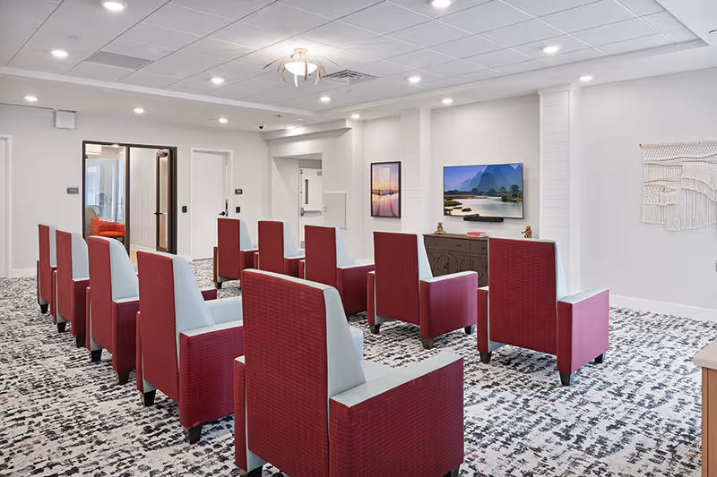 Bright community media room with rows of red and gray armchairs facing a wall-mounted TV and framed art.