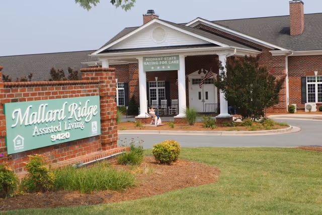 Brick assisted living building entrance with columns and a green 'Mallard Ridge Assisted Living' sign in the landscaped foreground.