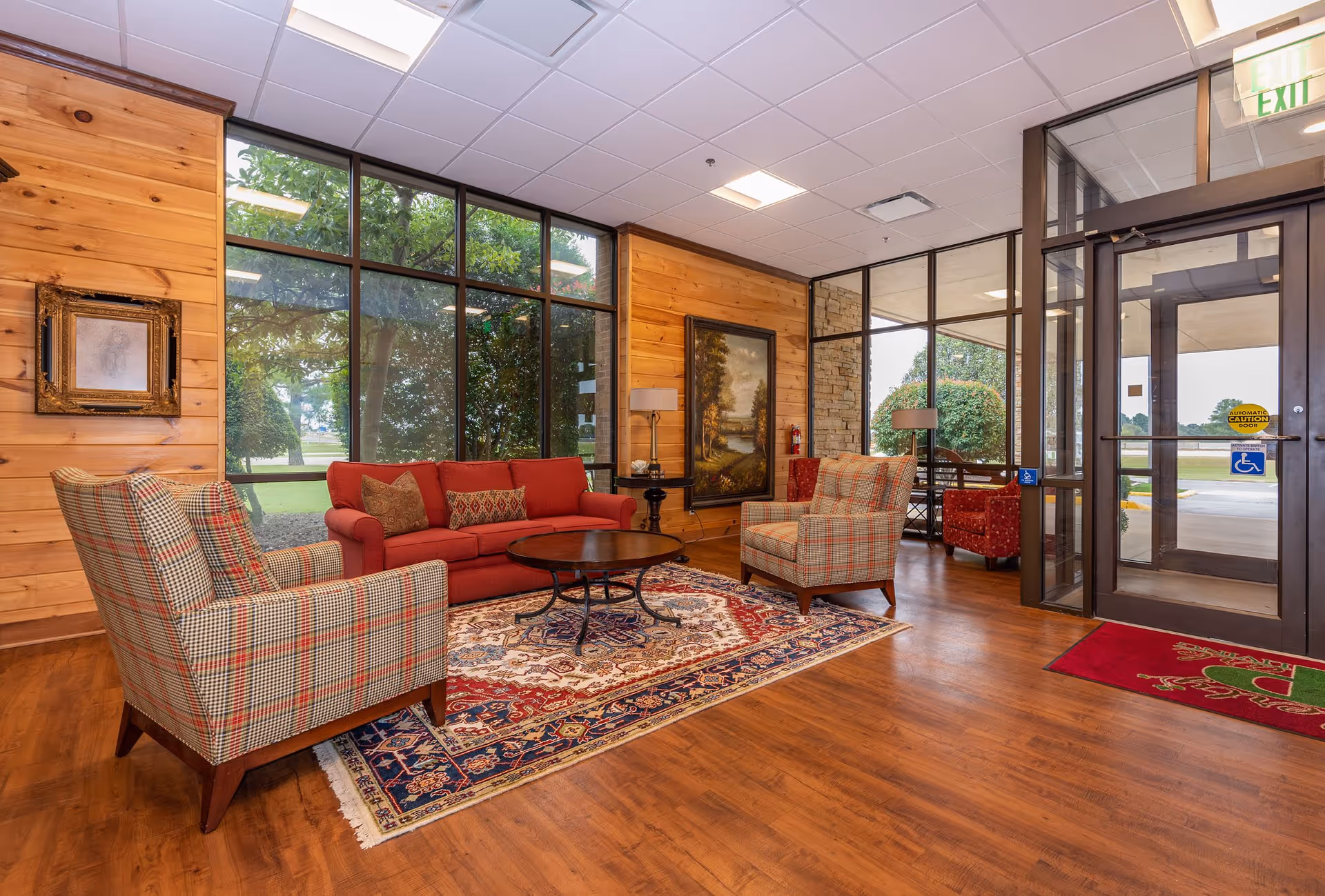 Well-lit assisted living lobby with a red sofa, plaid armchairs, a patterned rug, large windows, wood-paneled walls and glass entrance doors.
