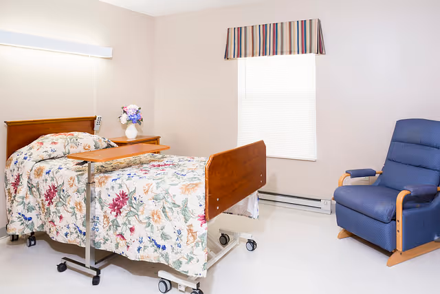A simple, clean bedroom in a senior living facility with a hospital-style bed covered in a floral bedspread, a wooden bedside table with a vase of flowers, a blue upholstered recliner chair, and a window with a striped valance.
