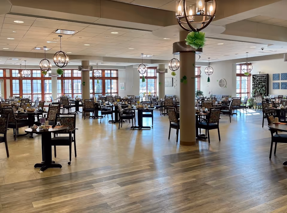 Spacious dining room with many set tables and chairs, large windows, and hanging light fixtures.