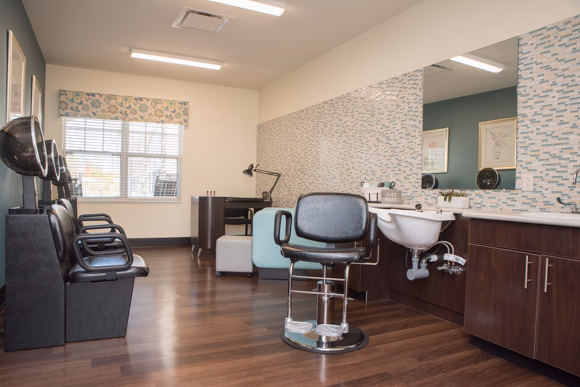 Interior view of a senior living facility hair salon with black salon chairs, hair dryers, a large mirror, a sink, and a desk with a lamp near a window with floral valance.