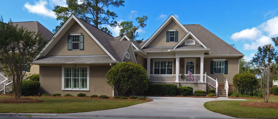 Front exterior of a single-story house with gabled roofs, a porch, driveway, and an American flag.