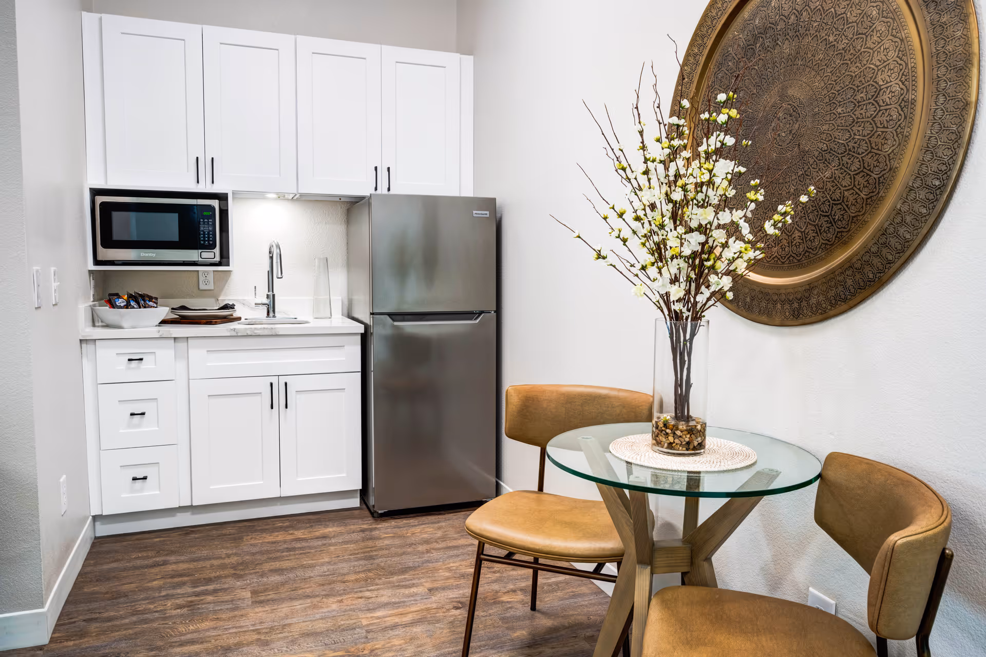 A small kitchen area with white cabinets, a stainless steel refrigerator, and a microwave above the sink. Next to the kitchen is a round glass table with a vase of white flowers and two brown cushioned chairs. A large decorative circular wall hanging is on the wall behind the table.