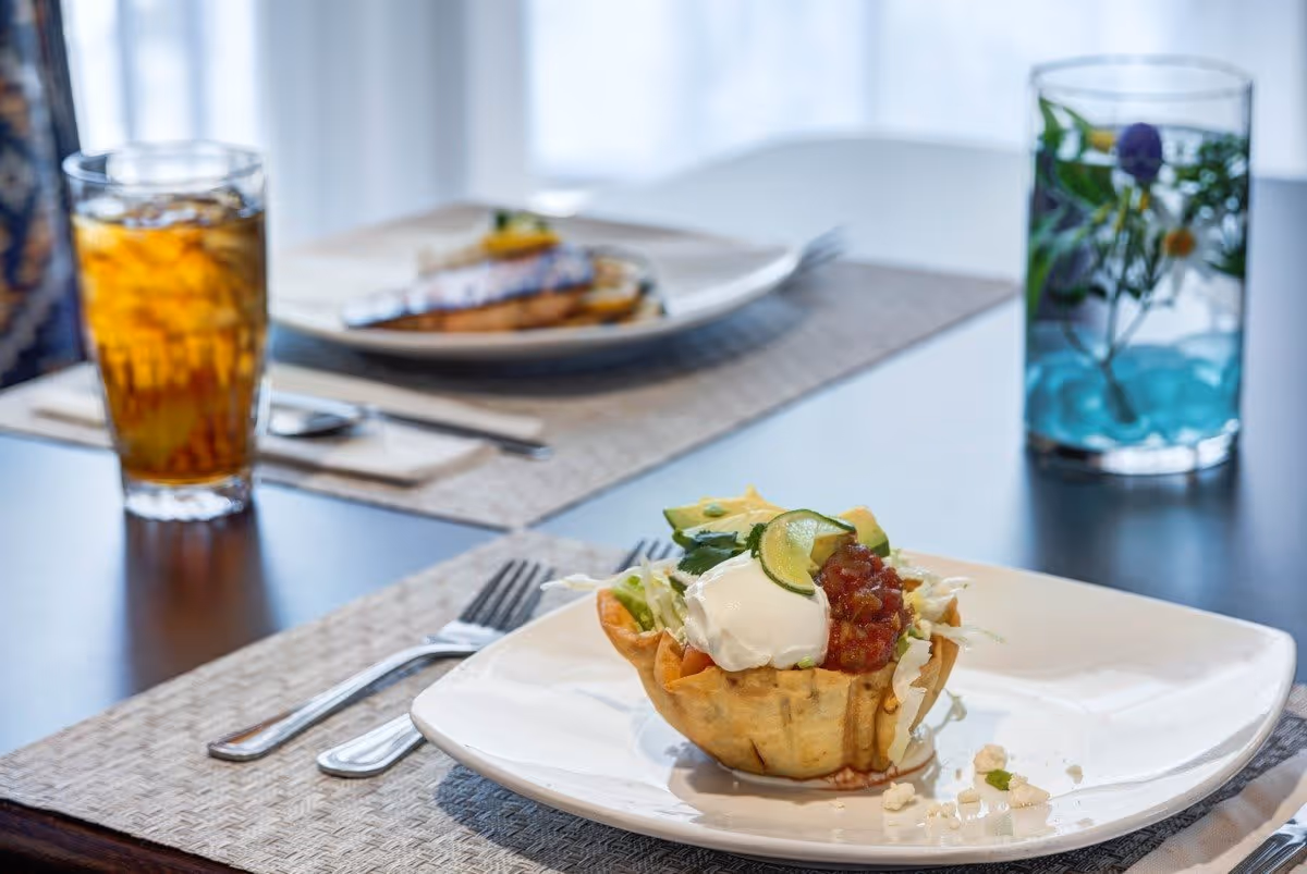 A close-up of a taco salad served in a crispy tortilla bowl on a white plate, garnished with sour cream, salsa, avocado slices, and lettuce. In the background, there is a glass of iced tea and another plate with food on a dining table set with placemats and utensils.