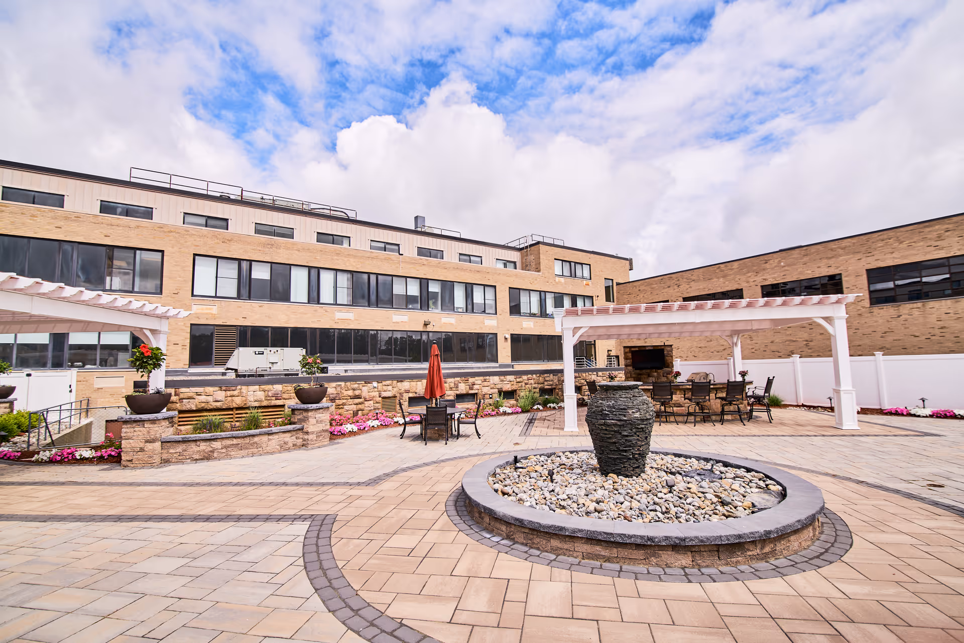 Outdoor courtyard area of CareOne at Concord featuring a circular stone water fountain surrounded by pebbles, paved walkways, flower beds with pink flowers, two white pergolas with seating and tables underneath, and a multi-story brick building in the background under a partly cloudy sky.