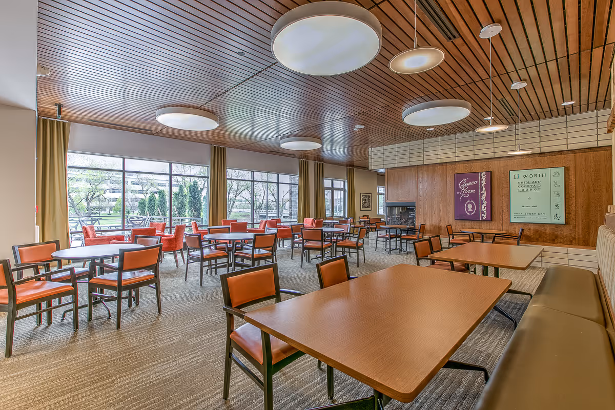A spacious dining room with multiple wooden tables and orange cushioned chairs. Large windows along one wall let in natural light and provide a view of greenery outside. The ceiling is wooden with modern circular light fixtures. There are two framed posters on a wood-paneled wall and a fireplace in the corner.
