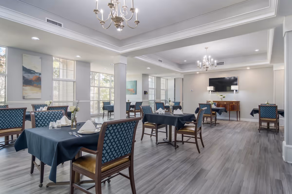 A spacious dining room with multiple tables covered in dark blue tablecloths, each set with plates, napkins, glasses, and silverware. The room features wooden chairs with blue patterned upholstery, large windows allowing natural light, light gray walls, and two chandeliers hanging from a tray ceiling. A flat-screen TV is mounted on the far wall above a wooden sideboard with two lamps and a small vase of flowers.