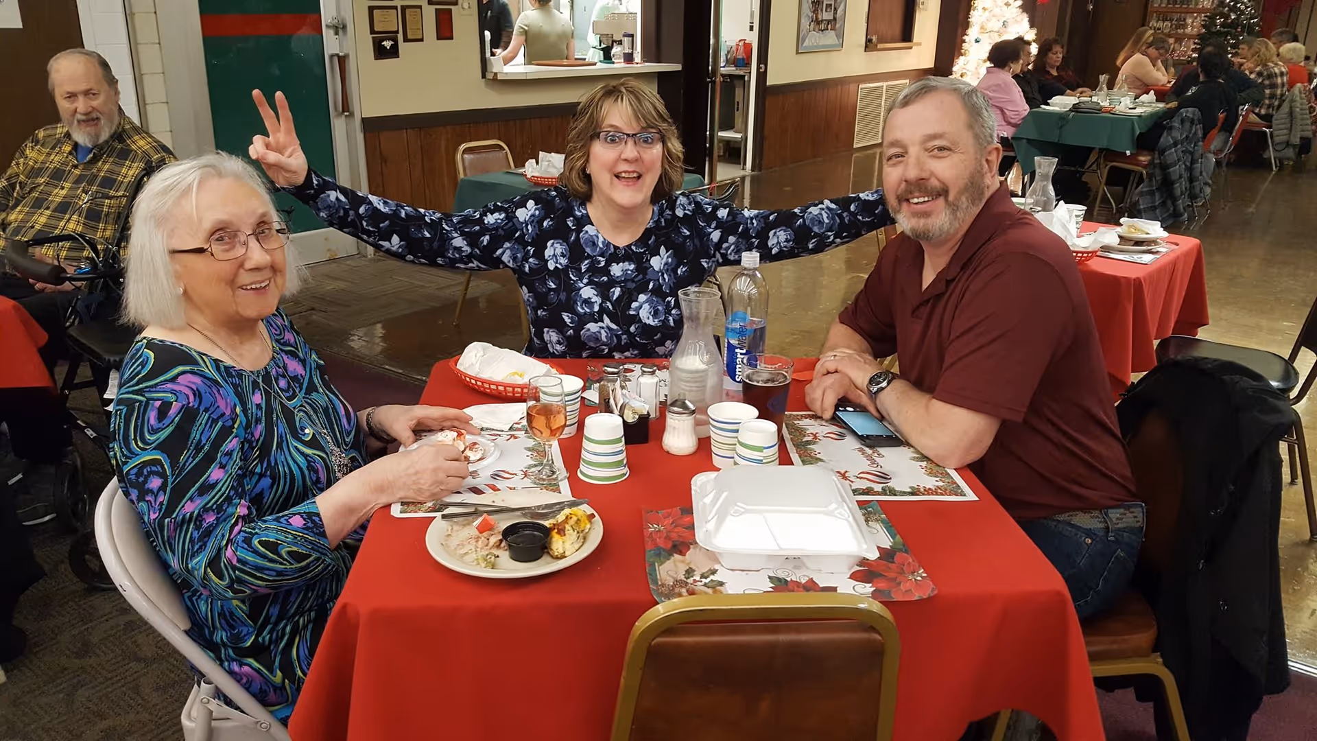 Three adults sitting at a table covered with a red tablecloth in a dining area. Two women and one man are smiling at the camera, with one woman playfully making a peace sign behind another's head. The table has plates with food, cups, and a water bottle. Other people are seated at tables in the background.