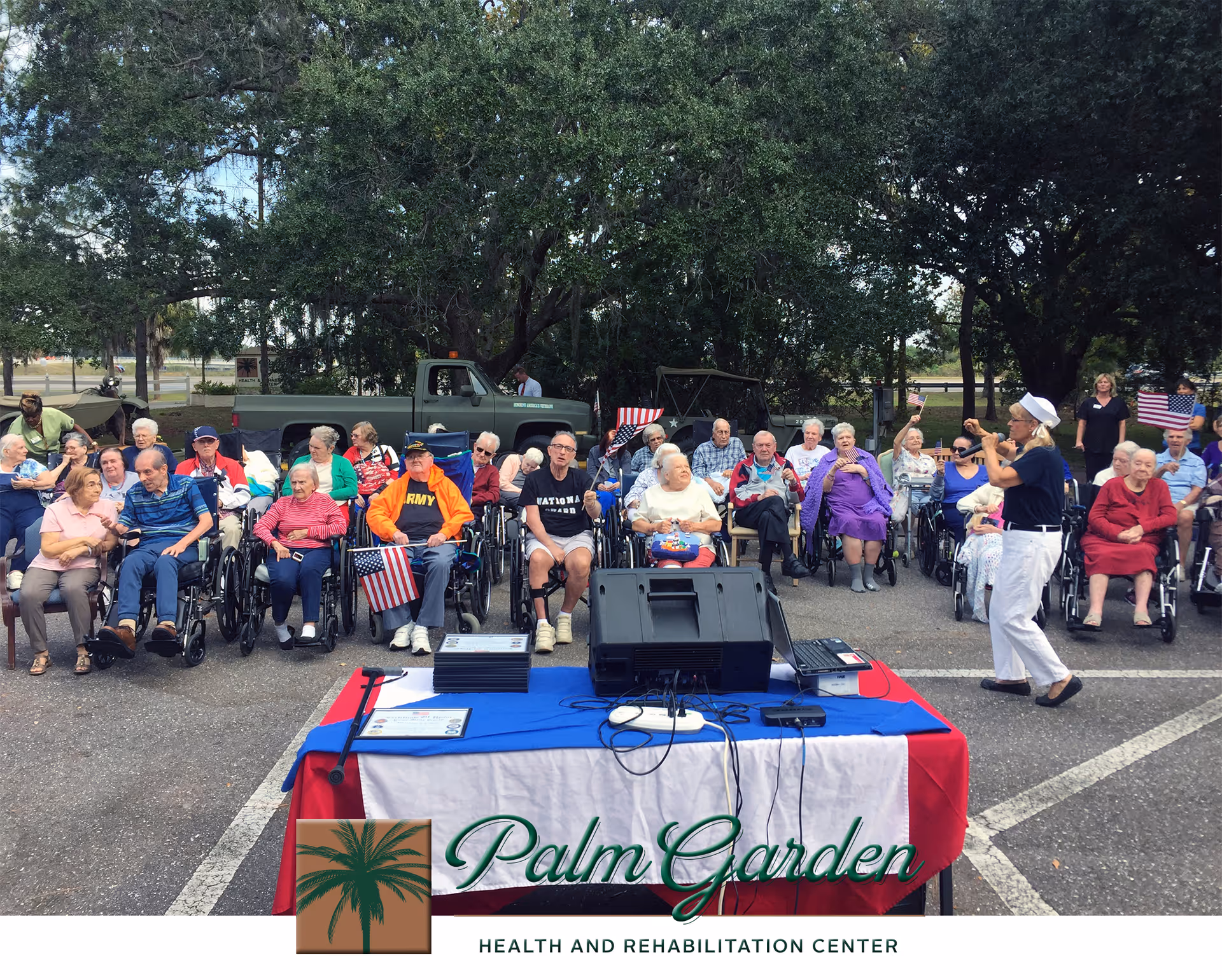 A group of elderly people, many in wheelchairs, seated outdoors in a parking lot area under large trees. A woman in white pants and a navy shirt is standing and speaking or singing into a microphone. Several people are holding small American flags. In the foreground, there is a table covered with a red, white, and blue cloth with audio equipment on it. Behind the group, there are two vintage military vehicles parked.