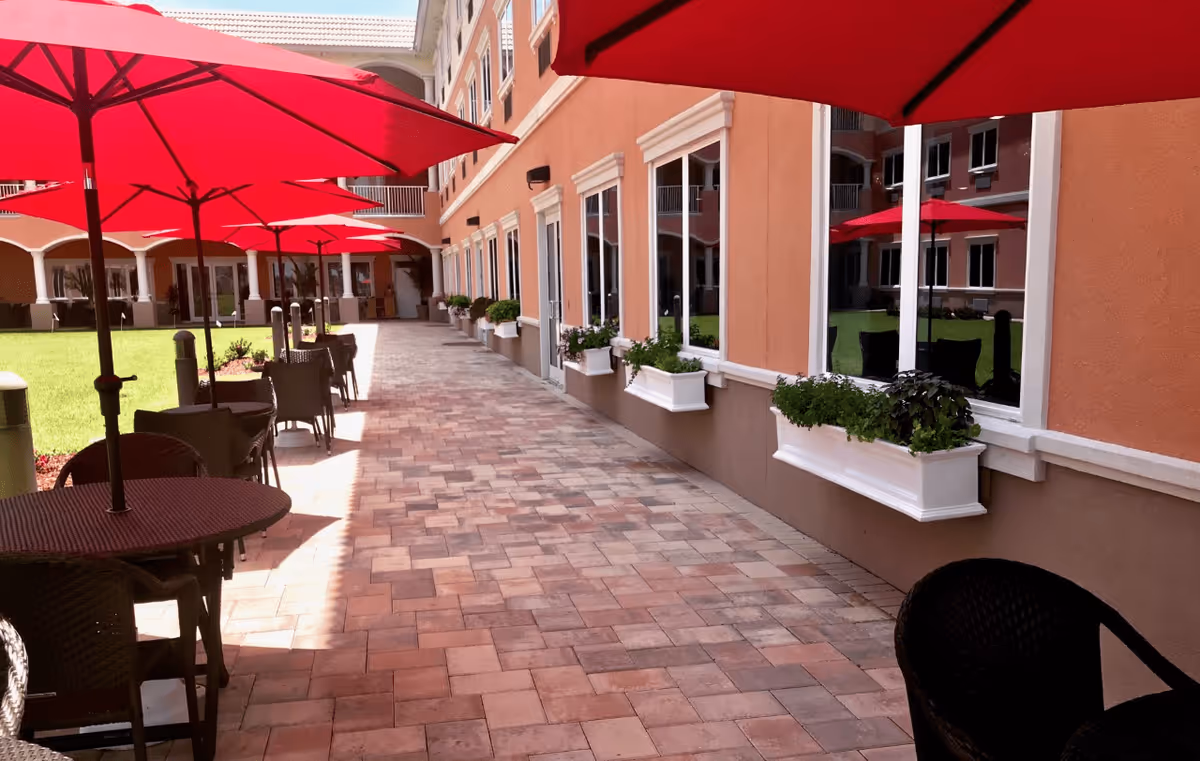 Outdoor patio area at Heritage Waterside Luxury Senior Living with red umbrellas shading round tables and chairs along a paved walkway next to a building with large windows and flower boxes.