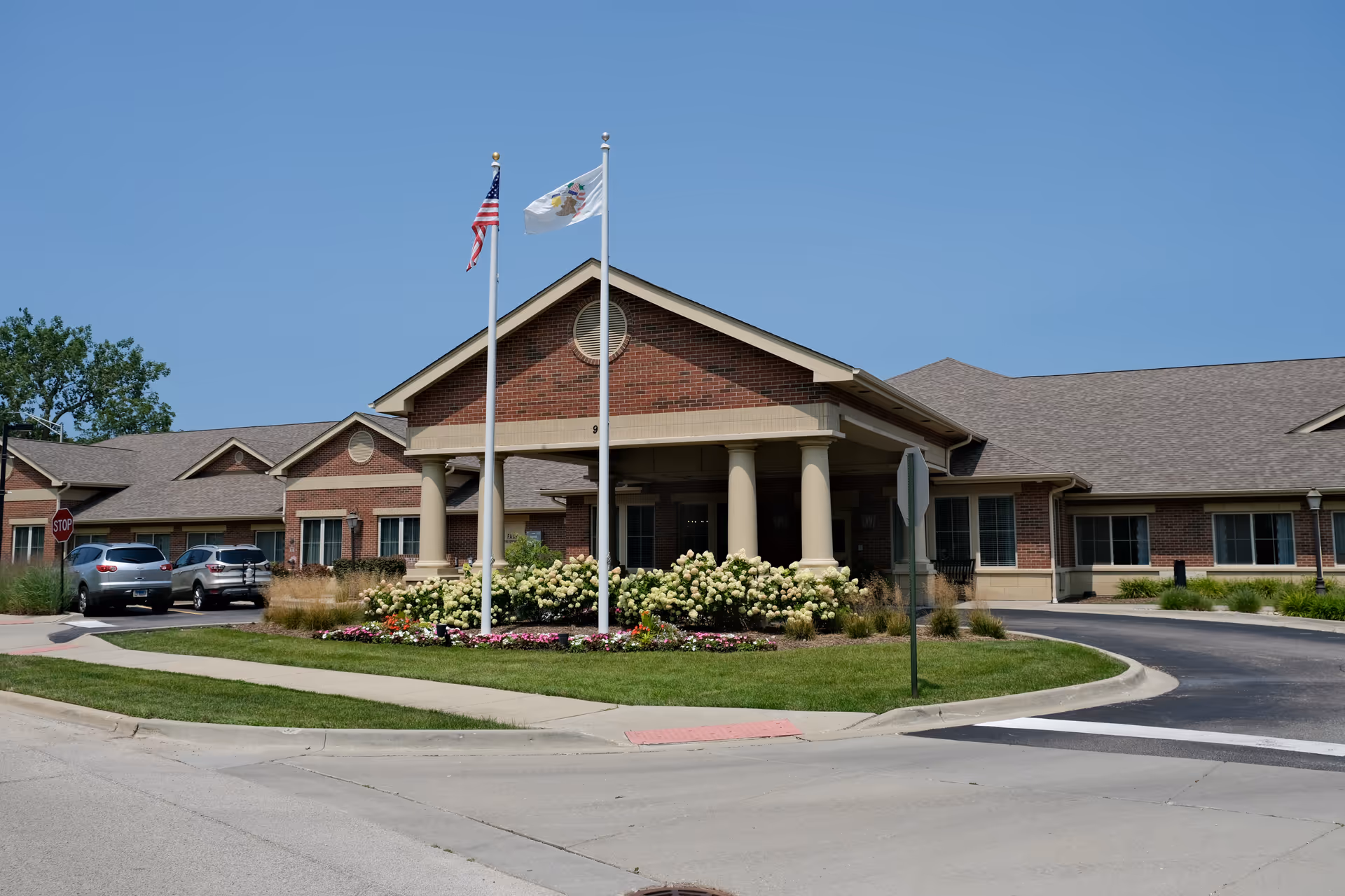 Brick single-story memory care building with a covered entrance, two flagpoles, and landscaped flower beds along a circular driveway.