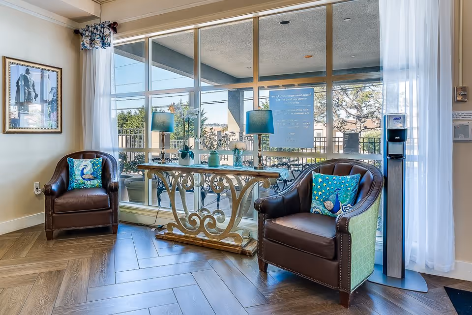 A cozy sitting area with two brown leather armchairs, each adorned with a decorative peacock-themed pillow. Between the chairs is an ornate wooden console table with two blue lamps and floral decorations. Large windows behind the table provide a view of an outdoor patio area with trees and a clear sky. White sheer curtains frame the windows, and a hand sanitizer dispenser is positioned near the right chair.