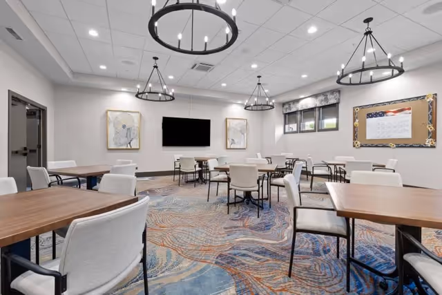 A spacious room with multiple wooden tables and cushioned chairs arranged neatly. The room has a patterned carpet with blue and orange swirls, three circular chandeliers hanging from the ceiling, a flat-screen TV mounted on the wall, two framed artworks on either side of the TV, and a bulletin board with an American flag design on the right wall. There are windows with floral valances letting in natural light.