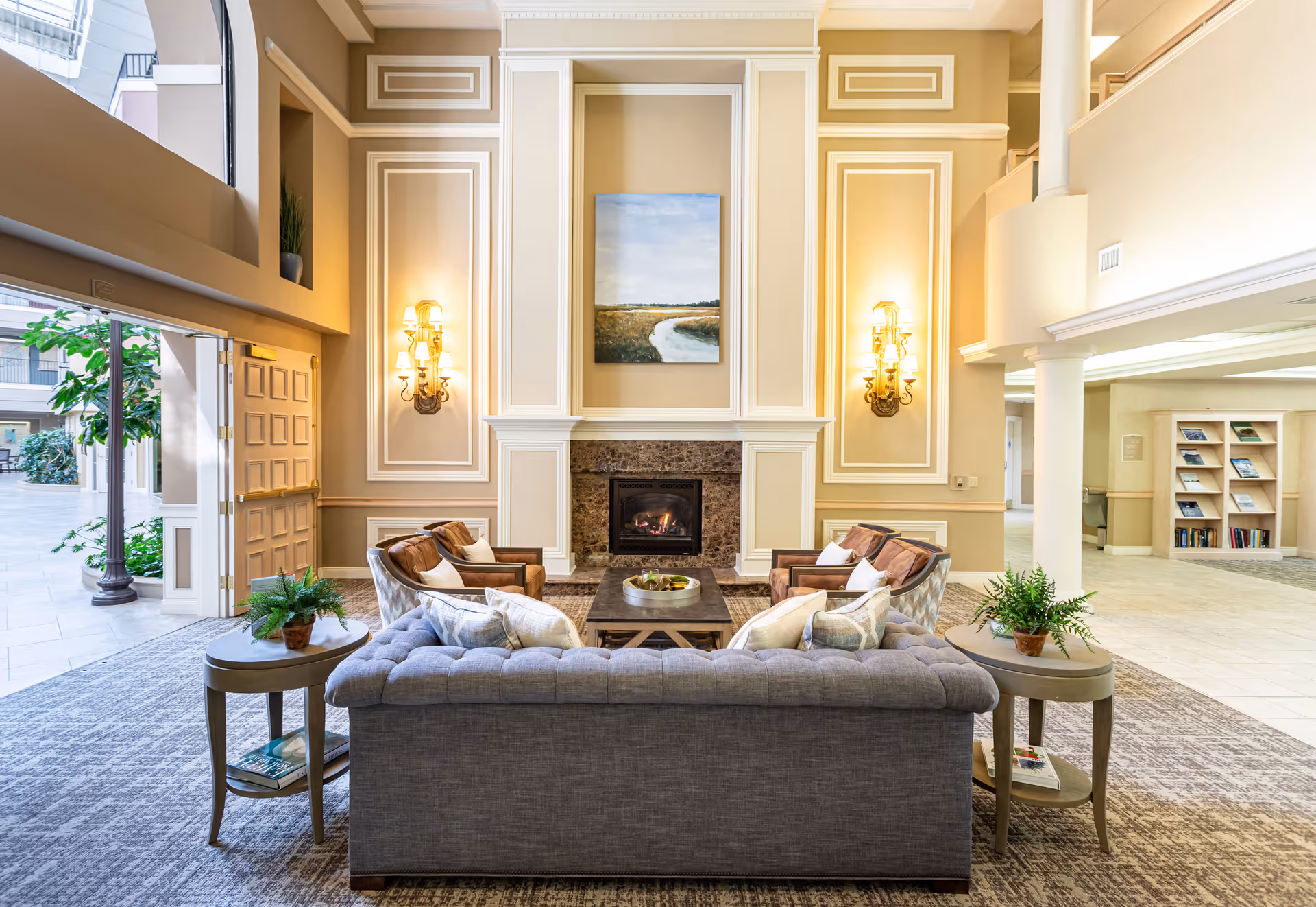 A bright and elegant living room area with a gray tufted sofa and four brown armchairs arranged around a wooden coffee table in front of a fireplace. The fireplace is framed by decorative molding and flanked by two wall sconces with lit lamps. Above the fireplace hangs a landscape painting. There are two small round side tables with potted plants on either side of the sofa. Large windows and an open door reveal an outdoor courtyard with greenery.