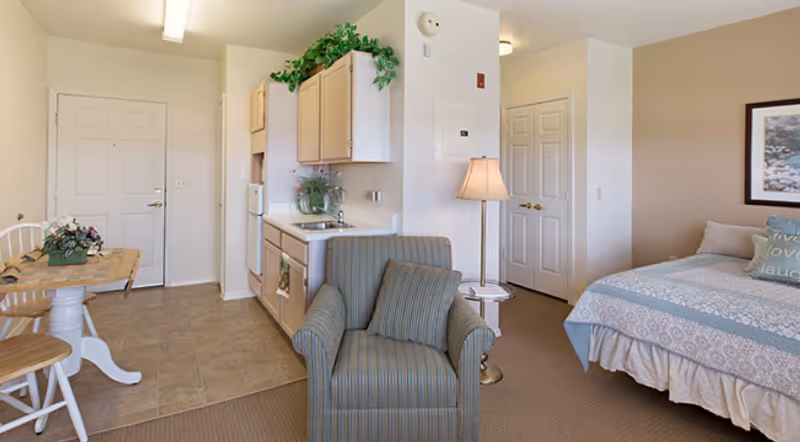 Studio-style assisted living interior showing a small kitchenette, armchair, dining table, and a bed in one open room.