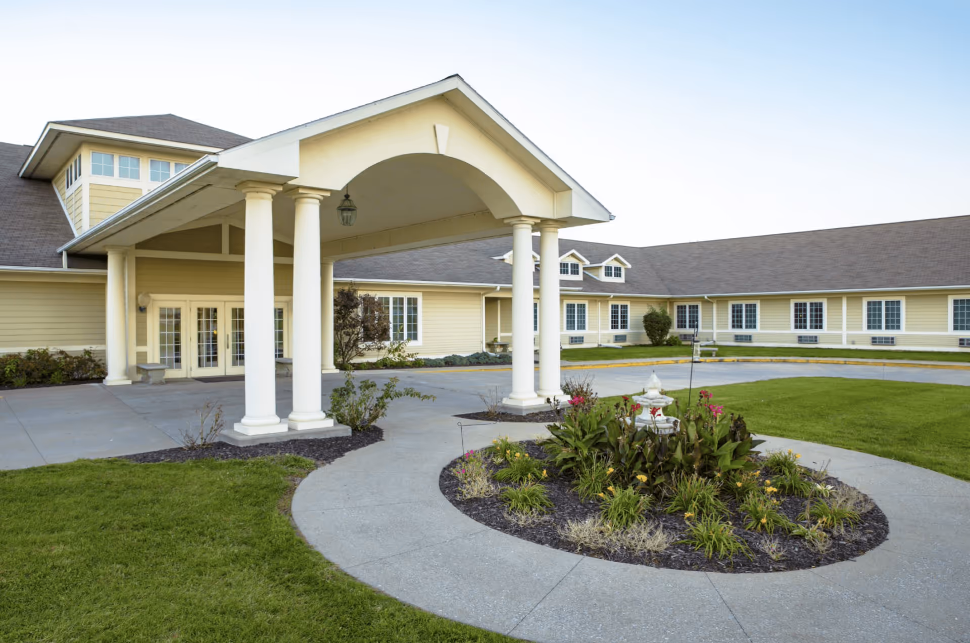 Exterior view of Homestead Of Centerville senior living facility showing a covered entrance with white columns, a circular driveway, landscaped garden with flowers and greenery, and a yellow building with multiple windows under a clear sky.