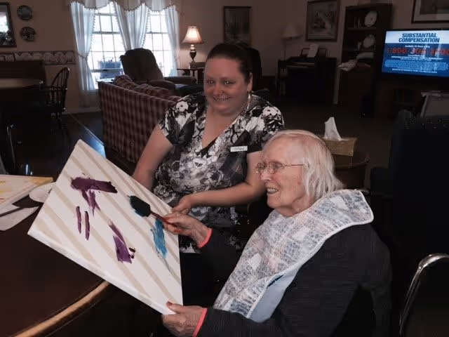 An elderly woman seated in a wheelchair is painting on a canvas with the assistance of a caregiver who is smiling and sitting beside her in a cozy living room setting with chairs, a table, and a television in the background.