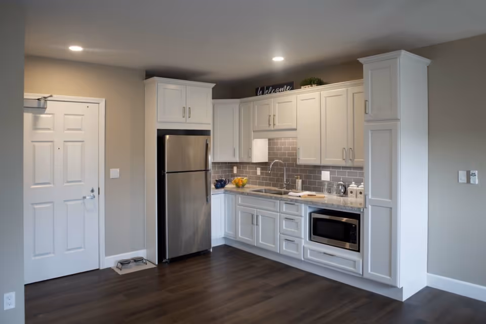 Modern kitchen area with white cabinets, a stainless steel refrigerator, a built-in microwave, a sink, and a gray tiled backsplash. The kitchen is well-lit with recessed ceiling lights and has dark wood flooring. There is a white door to the left and a small pet feeding station on the floor near the door.