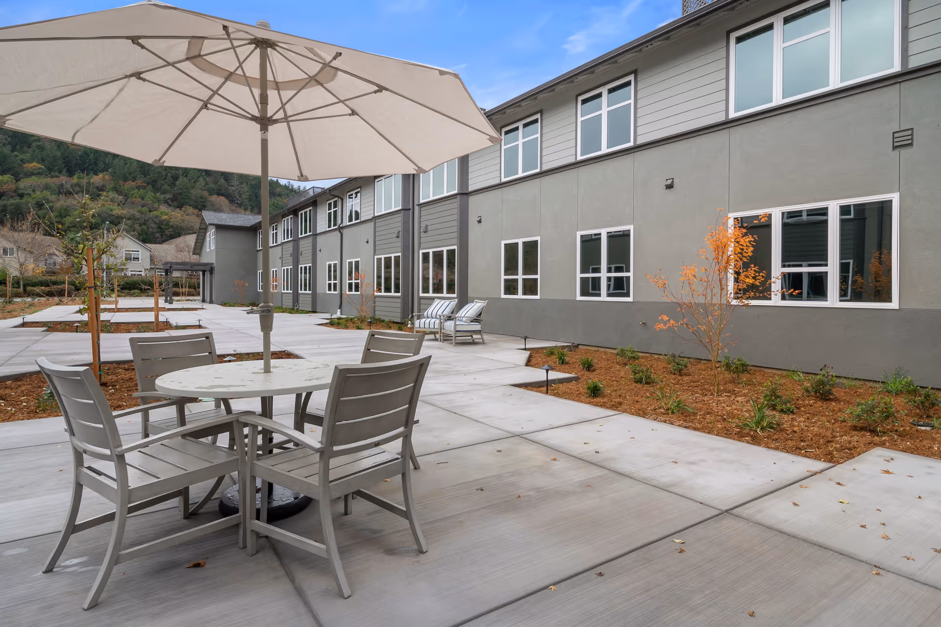 Outdoor patio area at Vine Ridge at Cloverdale featuring a round table with four chairs and a large umbrella. The patio is surrounded by a landscaped garden with small trees and shrubs, adjacent to a two-story building with multiple windows. Hills covered with trees are visible in the background under a clear blue sky.