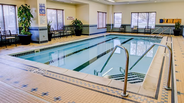 Indoor swimming pool with clear water, surrounded by tiled flooring and several chairs along the walls. Large windows allow natural light to enter the room, and there are potted plants placed near the seating areas.
