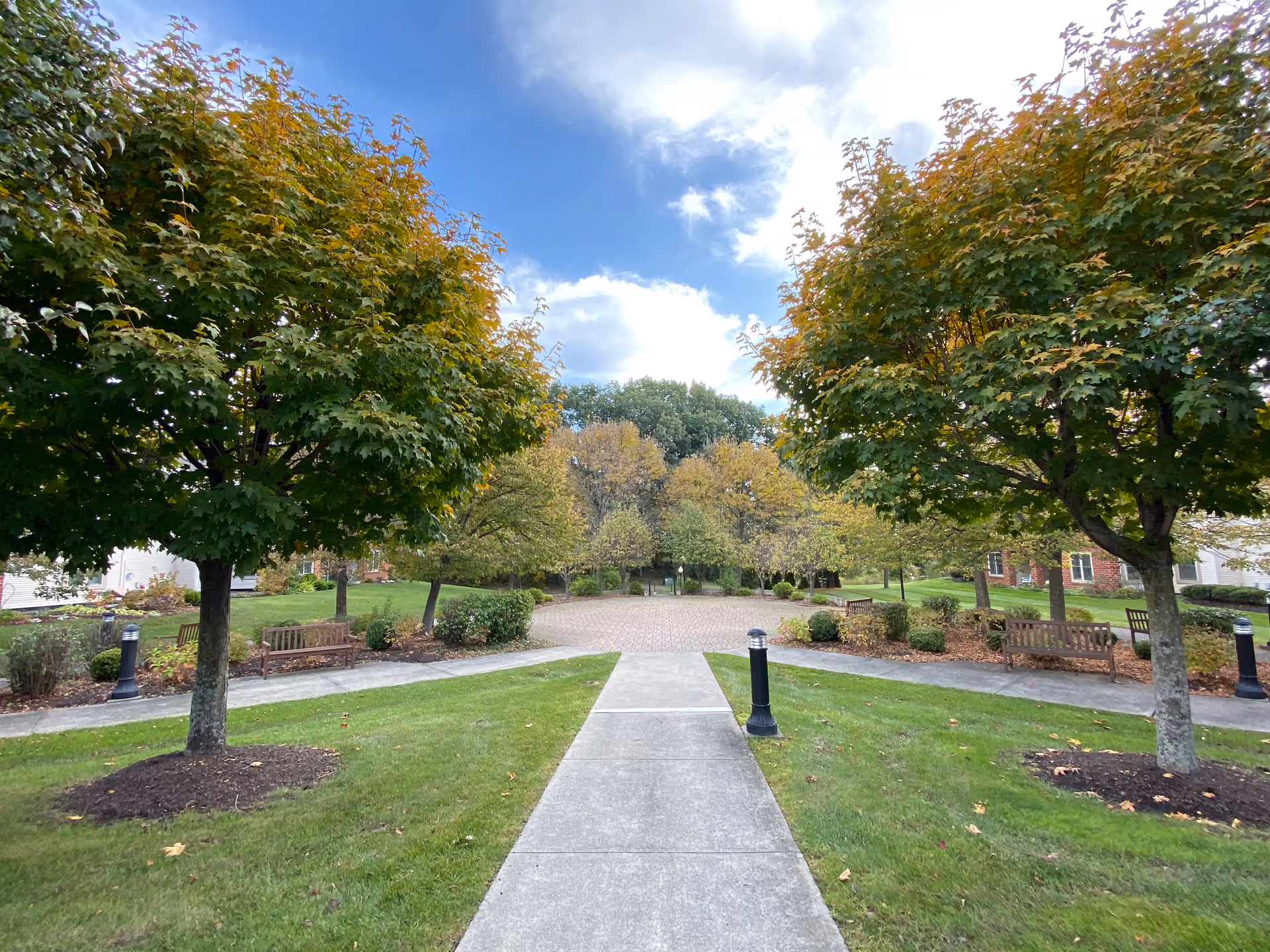 A paved walkway leading through a landscaped outdoor area with green grass, trees with some autumn foliage, benches, and lamp posts under a partly cloudy blue sky.