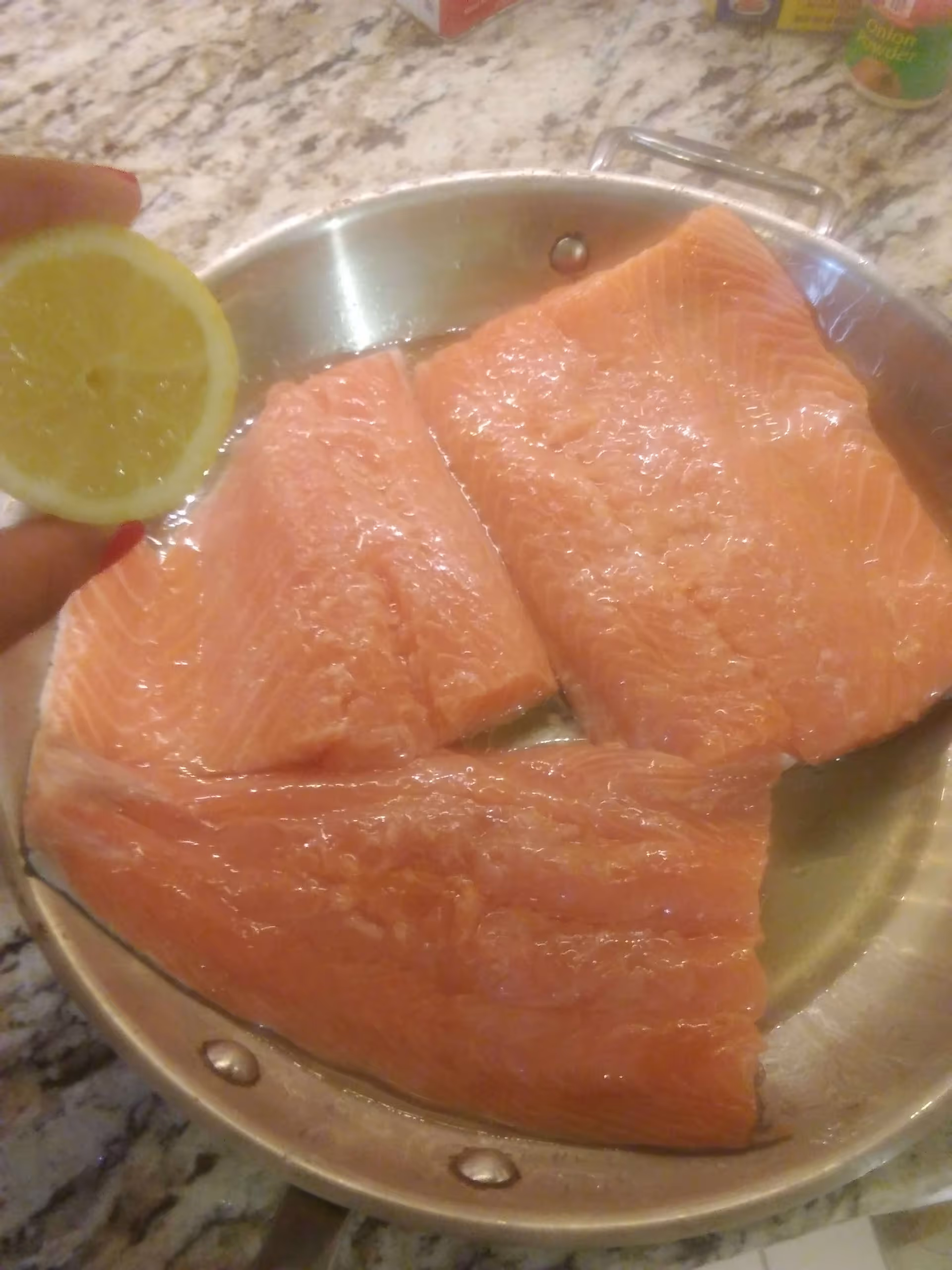Three raw salmon fillets in a metal pan on a granite countertop with a hand holding half a lemon above the pan.