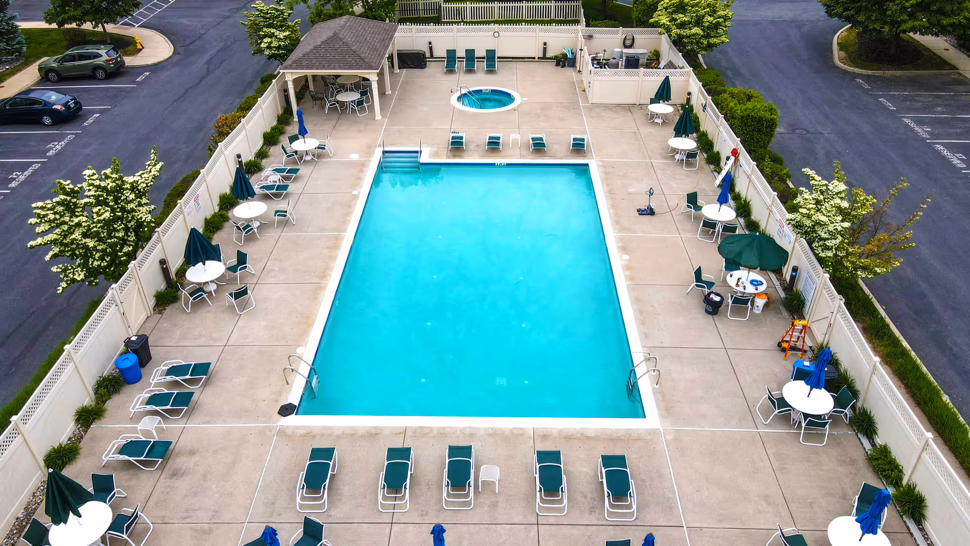 Aerial view of a rectangular outdoor swimming pool surrounded by lounge chairs, tables with umbrellas, and a small hot tub at one end. The pool area is enclosed by a white fence and there is a small covered seating area in one corner. The surrounding area includes parking spaces and greenery.