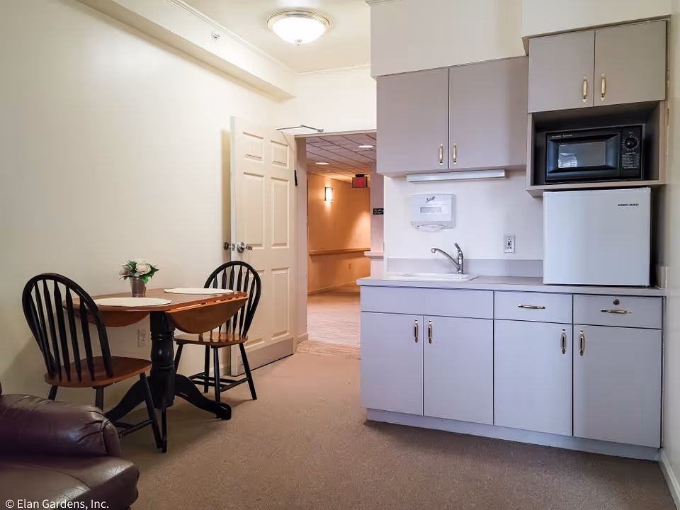 Small kitchenette with cabinets, sink, microwave and mini-fridge beside a small dining table and open door to a hallway.