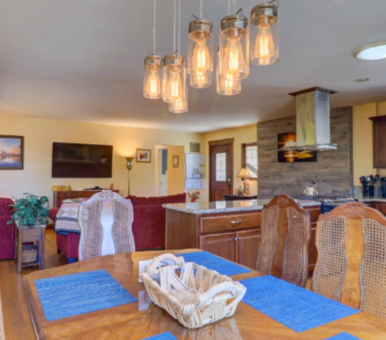 Interior view of a senior living facility showing a dining area with a wooden table, blue placemats, and a wicker basket centerpiece. Behind the dining area is a kitchen with wooden cabinets, a granite countertop island, and a stainless steel range hood. In the background, there is a living room with red sofas, a flat-screen TV mounted on the wall, a floor lamp, and framed pictures. The room is warmly lit with hanging pendant lights above the dining table.