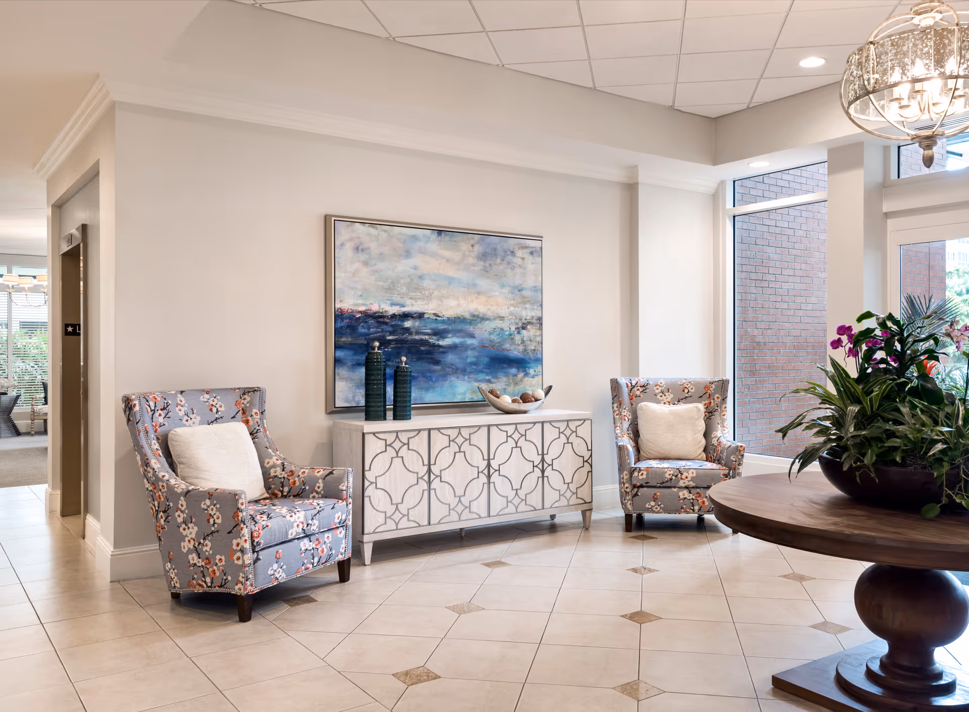 A bright and welcoming interior seating area with two floral-patterned armchairs each with a white cushion, a decorative white cabinet with a blue and white abstract painting above it, and a round wooden table with a large plant arrangement. Large windows let in natural light and a chandelier hangs from the ceiling.