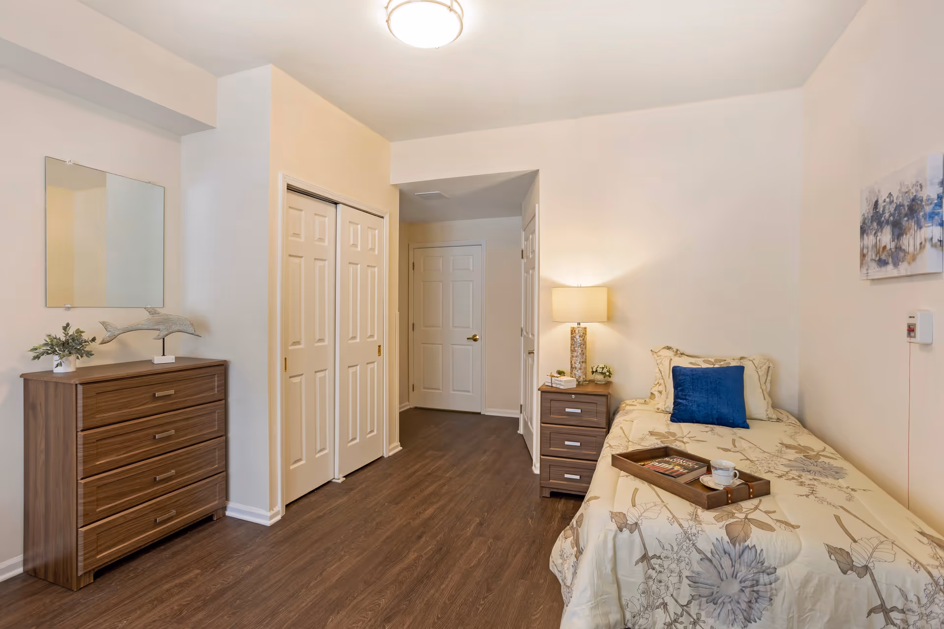 A neatly arranged bedroom with a single bed covered in a floral bedspread and a blue pillow. A wooden nightstand with a lamp, small plants, and books is beside the bed. Across from the bed is a wooden dresser with a decorative dolphin sculpture and a small plant. The room has light-colored walls, wood flooring, a ceiling light, and a hallway with white doors.